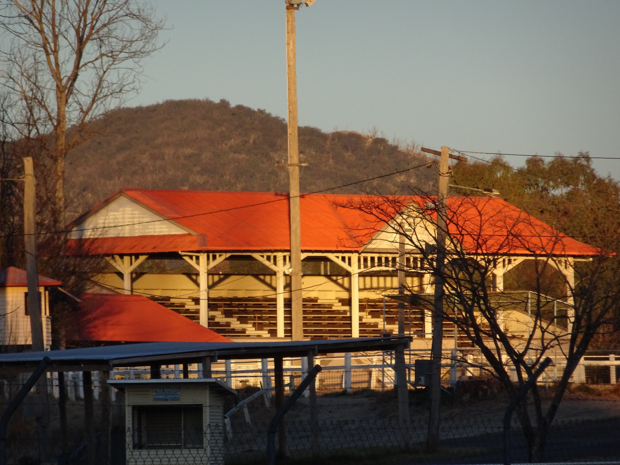 The showground grandstand with its red roof and white timber posts, lit gold at sunset with the hill behind